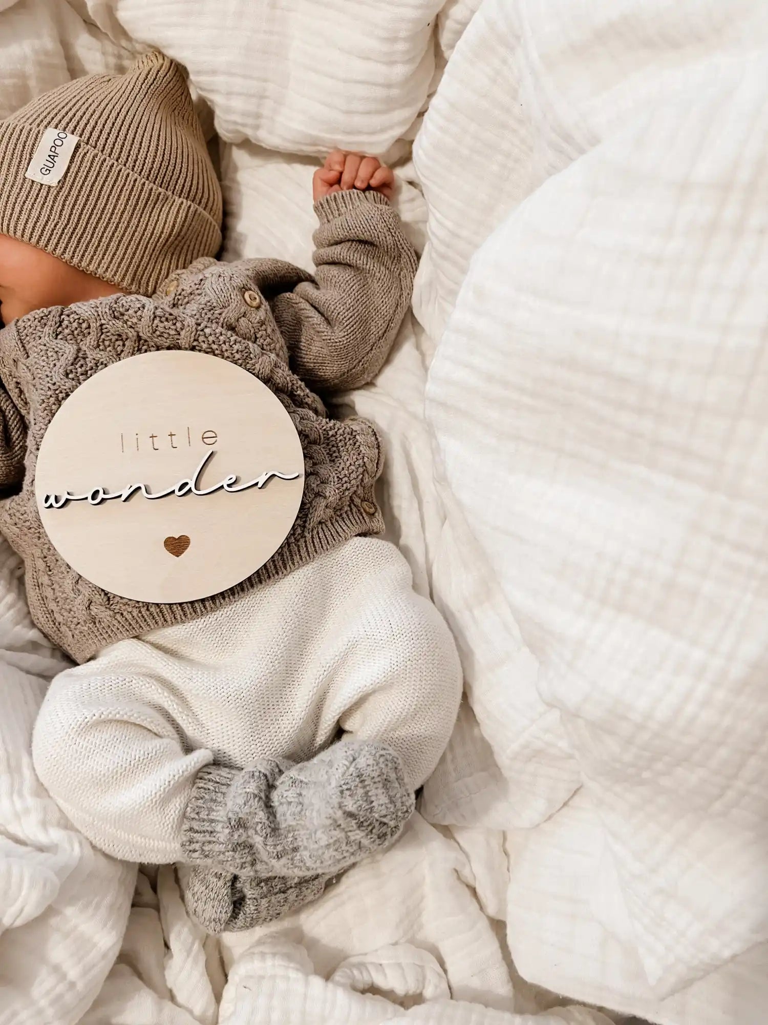 Ein Baby, das einen braunen gestrickten Pullover, weiße Hosen und eine beige Mütze trägt und einen hellen Holzscheibe hält.